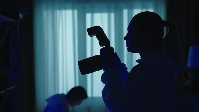 Forensic scientists collect evidence at the crime scene, in a dark apartment lit by blue red light from police sirens. A man inspects the murder weapon. A woman takes pictures with a photo camera.