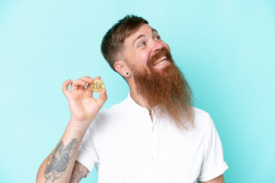 Redhead man with long beard holding a Bitcoin isolated on blue background looking up while smiling