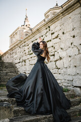 Fototapeta na wymiar Female in long black dress stands on stairs near ancient palace at sunset. Model girl in nature autumn day. Luxury woman near old Pidhirtsi Castle, Lviv region, Ukraine. Stylish bride. Back view