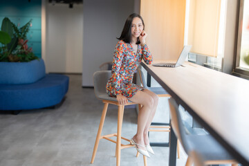 Asian female executive with short hair, company boss wearing a floral suit, sending a beautiful smile, having a laptop on the desk for business Sitting in a chair near the window in the office space