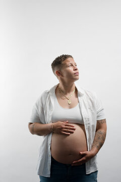 A pregnant woman shows off her big baby bump, protectively holding her pregnancy belly with both hands. Adding narrative, she is lesbian, married, and has a fresh, stylish butch aesthetic.