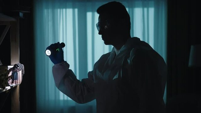 A young forensic expert in overalls and goggles is using a flashlight while examining a crime scene looking for clues. Medical examiner in a dark apartment lit by red and blue police sirens.