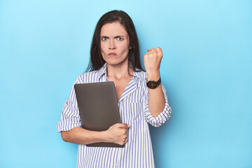 Young businesswoman holding laptop on blue showing fist to camera, aggressive facial expression.