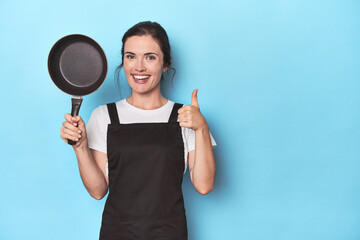 Woman with apron and pan on blue background smiling and raising thumb up