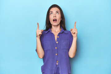 Young caucasian woman on blue backdrop pointing upside with opened mouth.