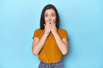 Young caucasian woman on blue backdrop shocked, covering mouth with hands, anxious to discover...