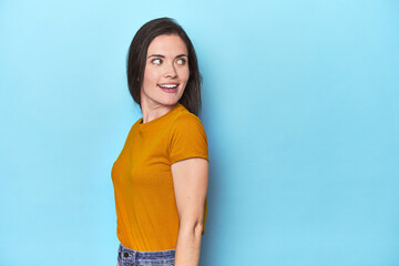 Young caucasian woman on blue backdrop looks aside smiling, cheerful and pleasant.