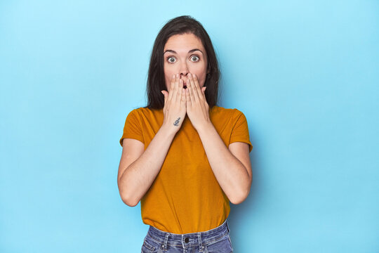 Young caucasian woman on blue backdrop shocked, covering mouth with hands, anxious to discover something new.