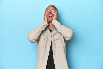 Middle-aged caucasian man on blue backdrop laughs joyfully keeping hands on head. Happiness concept.