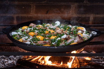 A large cast-iron skillet with fried meat and yellow eggs is cooked over an open fire.