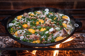 A large cast-iron skillet with fried meat and yellow eggs is cooked over an open fire.