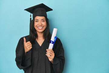 Filipina graduate with diploma on blue studio smiling and raising thumb up
