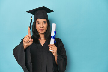 Filipina graduate with diploma on blue studio showing number one with finger.