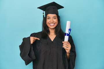 Filipina graduate with diploma on blue studio person pointing by hand to a shirt copy space, proud...