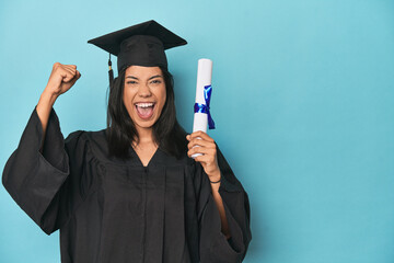 Filipina graduate with diploma on blue studio raising fist after a victory, winner concept.