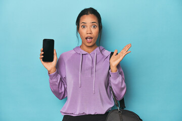 Filipina ready for gym with phone on blue surprised and shocked.