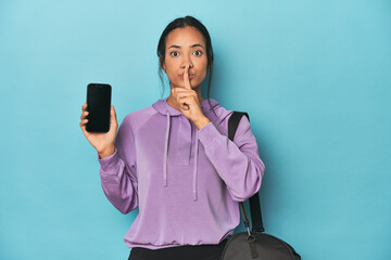 Filipina ready for gym with phone on blue keeping a secret or asking for silence.