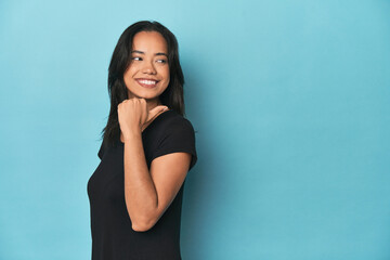 Filipina young woman on blue studio points with thumb finger away, laughing and carefree.