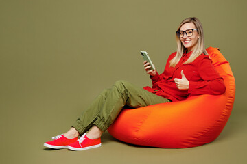 Full body side view young woman wear red shirt casual clothes glasses sit in bag chair use mobile...
