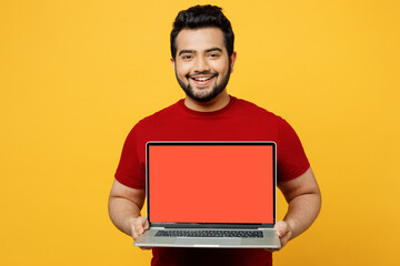 Satisfied smiling fun cool young happy IT Indian man wears red t-shirt casual clothes hold use work on laptop pc computer with blank screen workspace area isolated on plain yellow orange background.