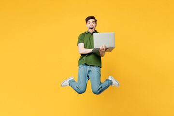 Full body surprised young shocked happy IT man wears green t-shirt casual clothes jump high hold use work on laptop pc computer isolated on plain yellow background studio portrait. Lifestyle concept.