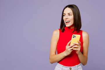 Young positive smiling happy fun woman she wears red tank shirt casual clothes hold in hand use mobile cell phone look aside on area isolated on plain pastel light purple background studio portrait.