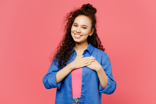Young smiling fun happy woman of African American ethnicity she wear blue shirt casual clothes put folded hands on heart look camera isolated on plain pastel pink background studio. Lifestyle concept.