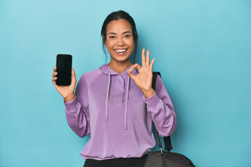Filipina ready for gym with phone on blue cheerful and confident showing ok gesture.