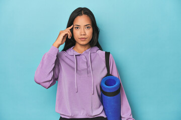 Filipina athlete with yoga mat on blue pointing temple with finger, thinking, focused on a task.