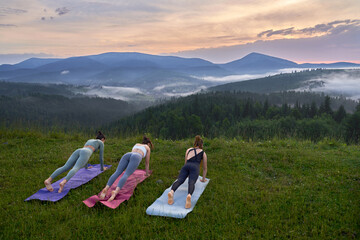 Naklejka premium Active young women in sport clothes doing yoga exercises on fresh air. Three fit ladies enjoying outdoors training during summer sunset.