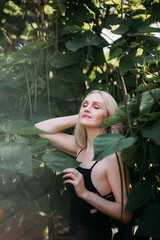 A positive pretty woman of age with long blond hair, in a black long sexy dress, stands and relaxes among the bushes with large green leaves in the city park.