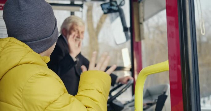 Side view of bus driver saying hello to passenger. Old man sitting, holding hand on helm, wearing suit greeting, waving to small boy. Concept of routine and urban lifestyle.