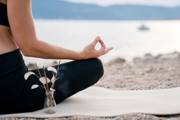 Meditation on sea beach with incense sticks. Woman practicing hatha yoga. Girl relaxing in wellness trip, retreat in nature outdoors. Concept of mental health, calmness, mindfulness, female wellbeing
