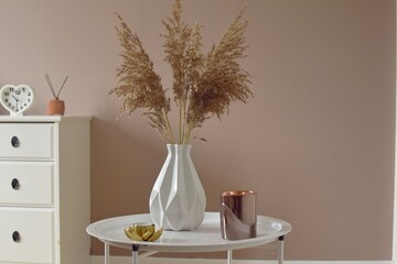 On a round, white table there is a vase with dry flowers, next to candle
