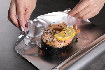 Woman wrapping tasty salmon with lemon and spices in aluminum foil at grey textured table, closeup