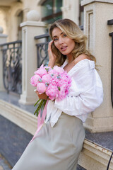 Elegant attractive blond woman holding amazing pink bouquet with pions.  Walking  outdoor in european city.  Wearing casual summer outfit, white blouse, silk skirt.