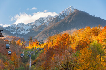 Autumn landscape with mountain range green, yellow red foliage of forest trees illuminated by the sun. High mountains with snow capped peaks with cable car, and passenger cabins rising up.