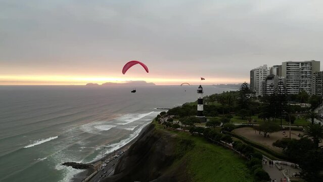 Aerial travelling shot showing a pair of paragliders flying in the coast near a lighthouse at sunset time in the middle of a park next to some buildings with the ocean and an island in the background