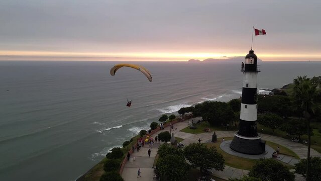 Aerial pan shot shows a paraglider flying at sunset time passing next to a lighthouse and turning when are people walking in a boulevard below him