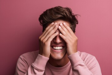 Lifestyle portrait photography of a happy boy in his 30s covering one eye against a dusty rose background. With generative AI technology