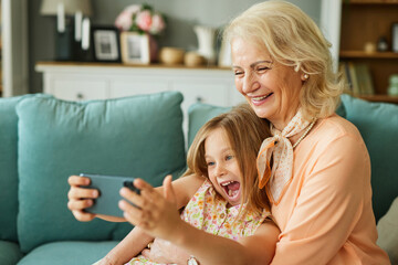 Happy grandmother and granddaughter taking selfie using smartphone in the living room - obrazy, fototapety, plakaty