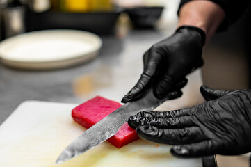 Fototapeta premium Close-up of hand sushi chef in gloves slices fresh tuna fish fillet at sushi bar.