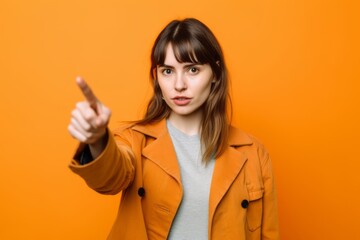 Headshot portrait photography of a glad girl in her 20s pointing up against a tangerine orange background. With generative AI technology