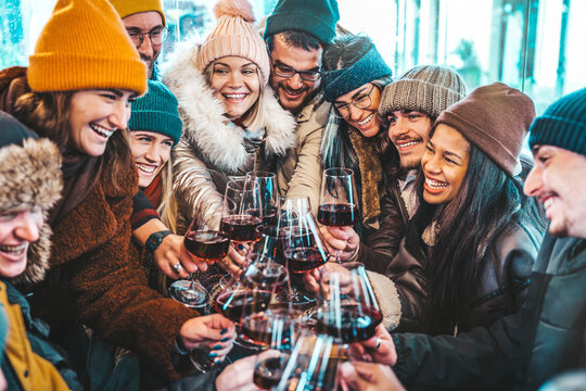 Happy multiracial friends toasting red wine at restaurant terrace - Group of young people wearing winter clothes having fun at outdoors winebar table - Dining life style and friendship concept