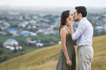 Man and woman in love stand on hill top kissing against breathtaking view. Concept of posing for romantic photoshoot on blurred town background