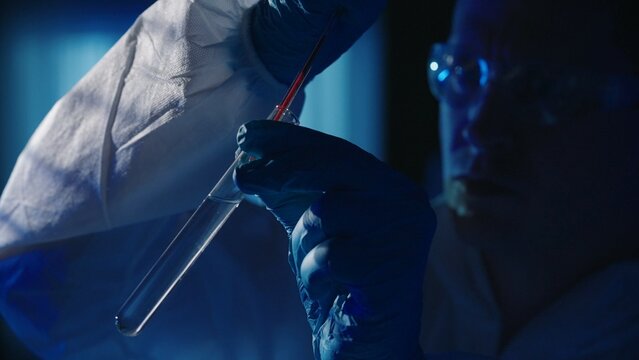 A forensic investigator adds a blood sample to a test tube with a reagent using a pipette. A man collects evidence at the crime scene, in a dark apartment lit by blue police sirens.