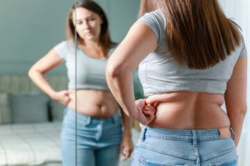 Displeased young woman touching her belly looking in mirror at home