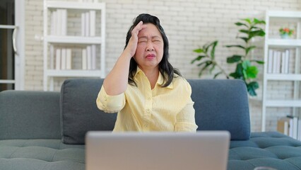 Asian elderly woman having headache migraine while working on laptop sitting in sofa at living room. Tired stressed upset senior woman having headache. Health problem concept