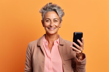 Medium shot portrait photography of a tender mature woman taking a selfie with his mobile against a pastel orange background. With generative AI technology