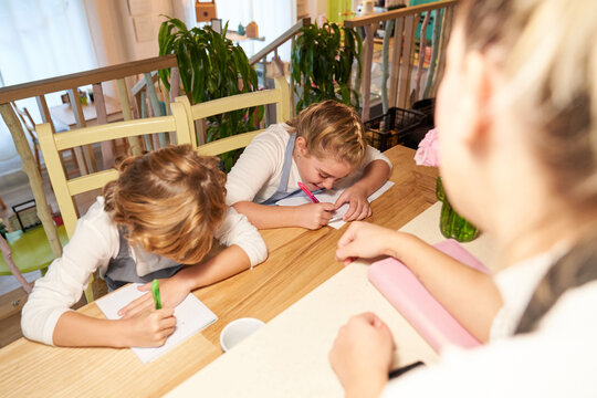 Girl in front of two children sitting in wooden chairs and with apron writing in the notebook - Powered by Adobe